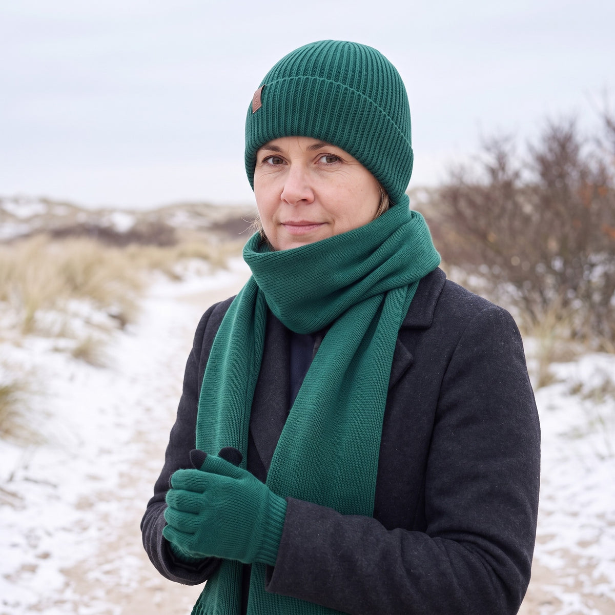 Vrouw met donkergroene gebreide sjaal met franjes en bijpassende muts en handschoenen op een besneeuwd duinpad
