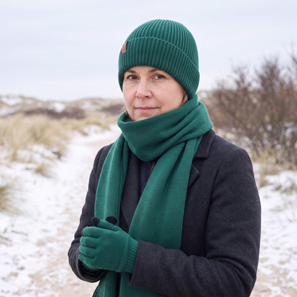 Vrouw met donkergroene gebreide sjaal met franjes en bijpassende muts en handschoenen op een besneeuwd duinpad